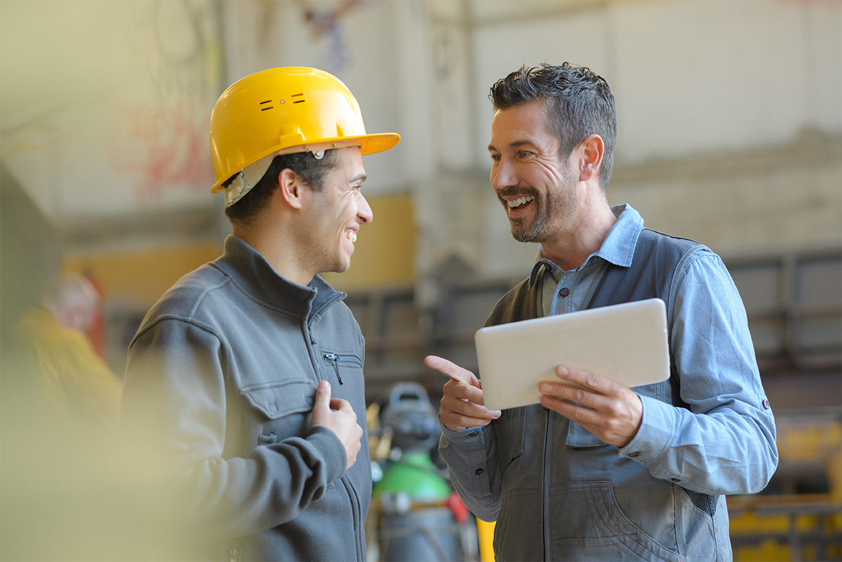 Two employees are talking to each other. One of them is wearing a protective helmet. The other holds a tablet in his hand. They are both smiling.